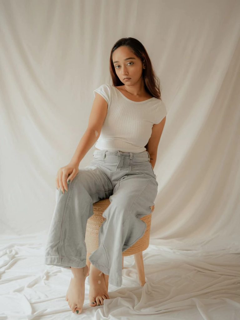 Meghna Ghosh seated in a minimalist studio setting wearing a white top and relaxed trousers